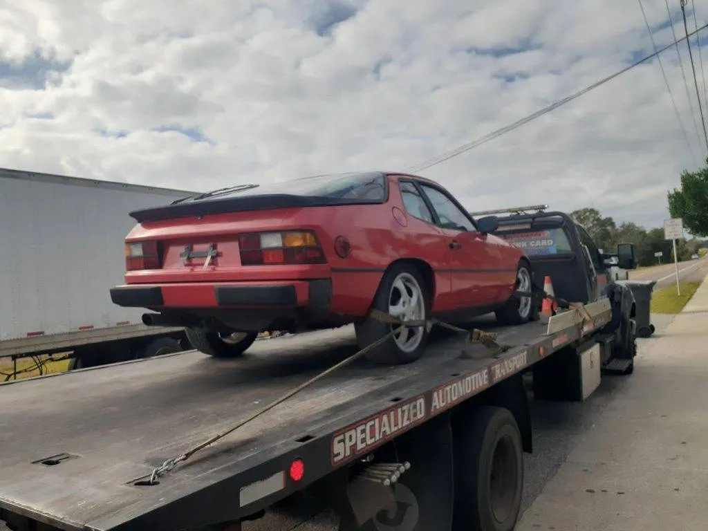 Red Porsche 944 coupe strapped down on a black flatbed beside a white semi trailer