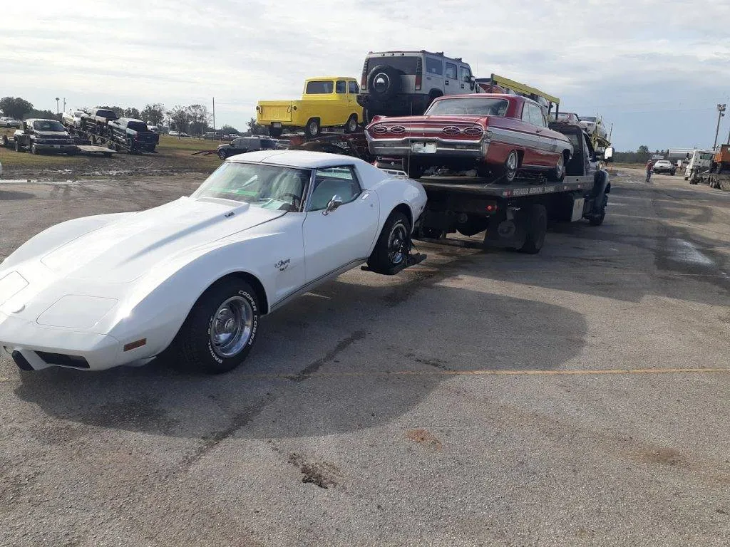 White Chevrolet Corvette Stingray parked in an open lot next to a loaded flatbed