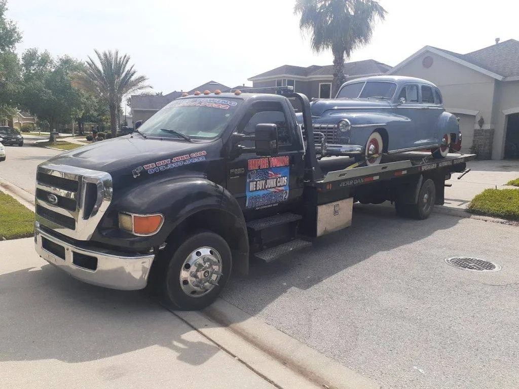 A blue vintage Cadillac coupe rides on an Empire Towing flatbed in a neighborhood