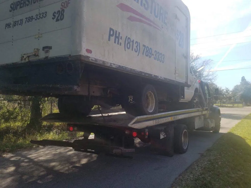 White box truck loaded on a flatbed heads along a tree lined road
