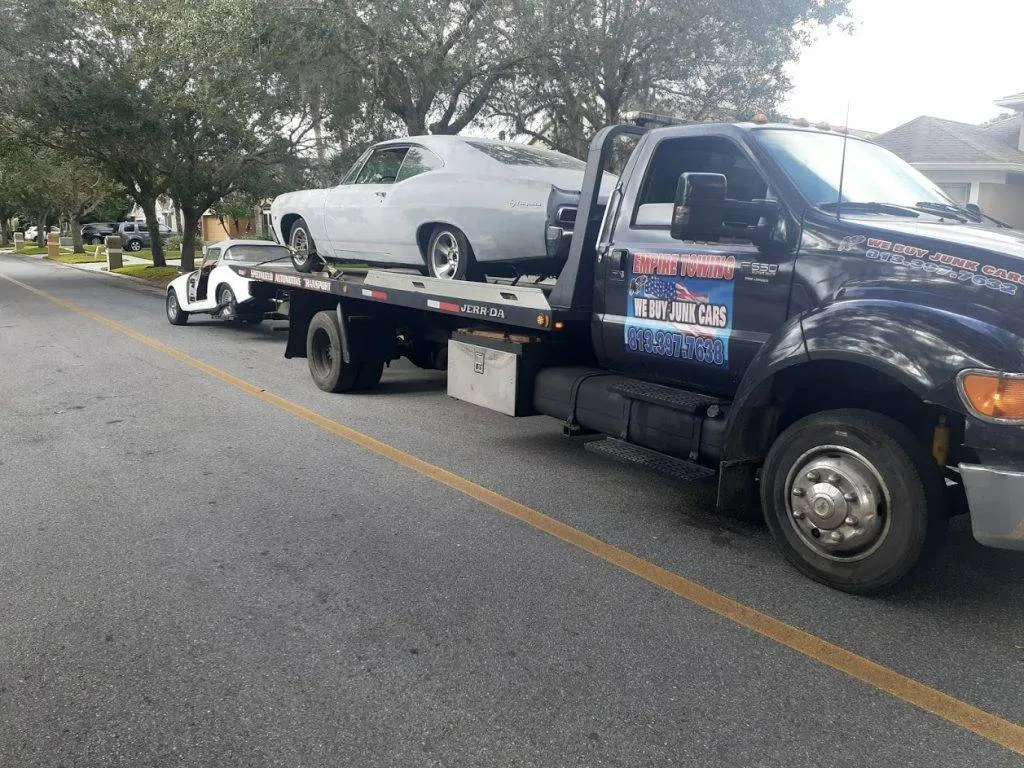 Along a leafy neighborhood street, a flatbed carries a white classic coupe