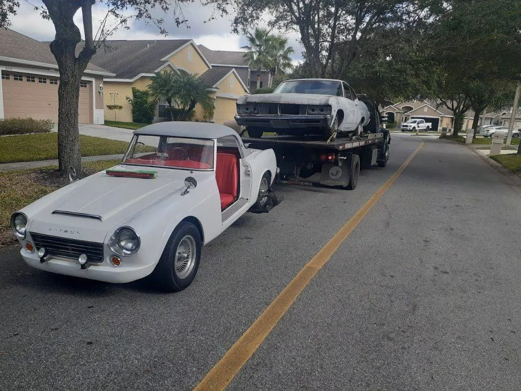 A vintage white Datsun roadster with red seats sits behind a loaded flatbed